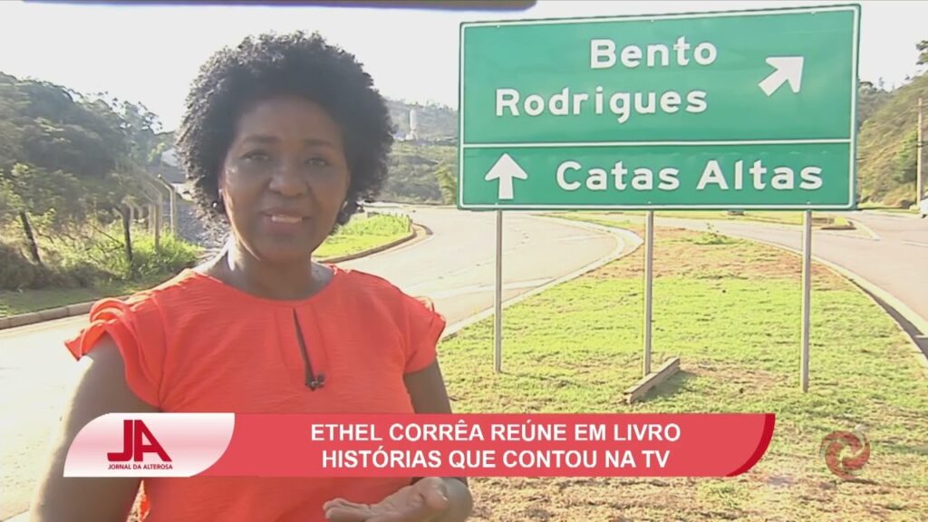 Ethel em uma rodovia, em frente a uma placa de transito que indica a diração para as cidades de Bento Rodrigues e Catas Altas. Usa uma blusa vermelha e cabelos soltos curtos. 