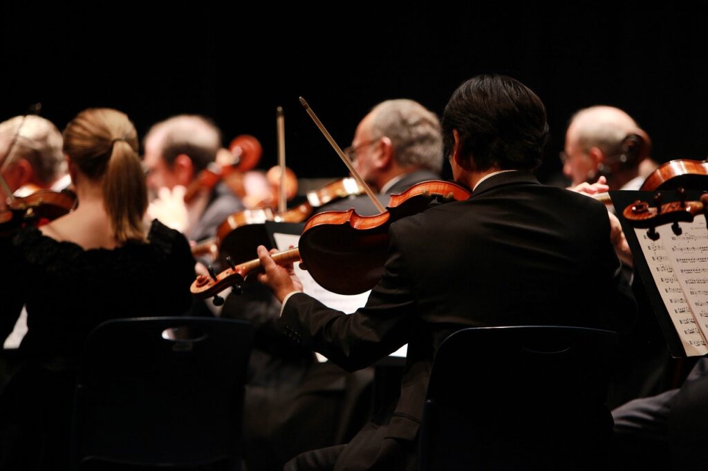 Pessoas de costas tocando instrumentos de orquestra. Todos usam preto e o fundo também, fazendo com que o destaque luminoso e colorido seja a faixa horizontal central da foto emoldurada pela predominância da cor preta