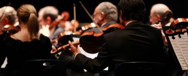 Pessoas de costas tocando instrumentos de orquestra. Todos usam preto e o fundo também, fazendo com que o destaque luminoso e colorido seja a faixa horizontal central da foto emoldurada pela predominância da cor preta