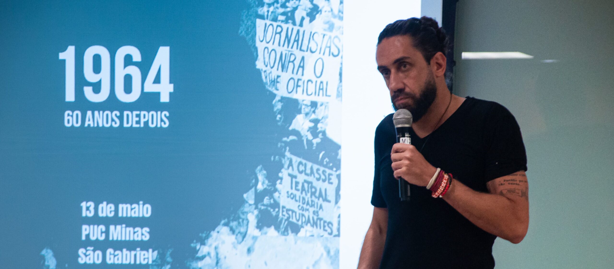 Homem branco usando uma camisa preta com um microfone na mão esquerda, em pé com um fundo com o nome da palestra do JFest