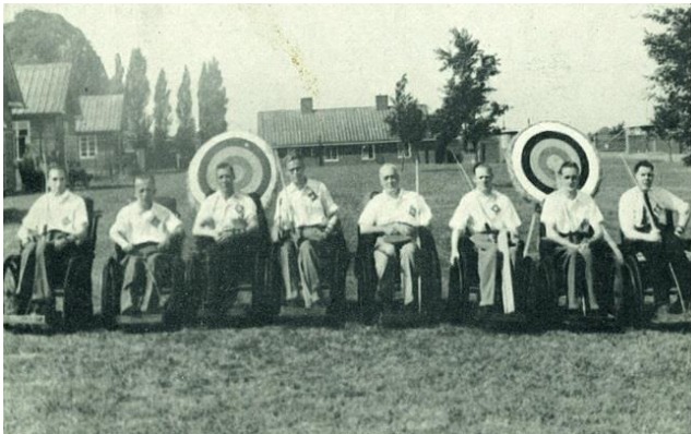 Oito homens em cadeiras de rodas em frente a alvos de arco e flecha. Foto em preto e branco.