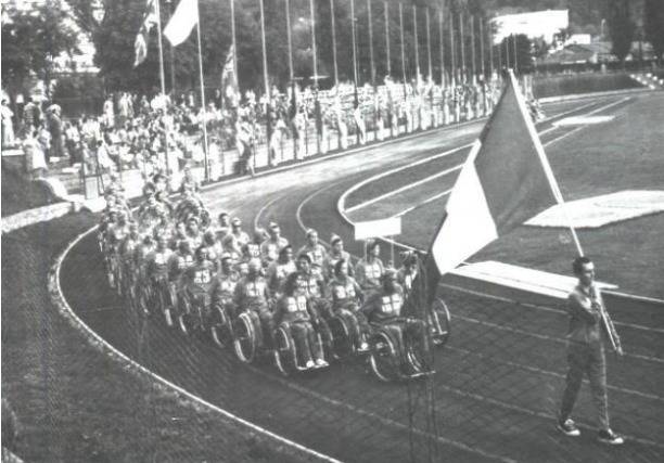 Foto em preto e branco. A foto se ambienta em uma pista de atletismo. Um homem com uma bandeira está a frente de uma fila de vários atletas em cadeiras de rodas.