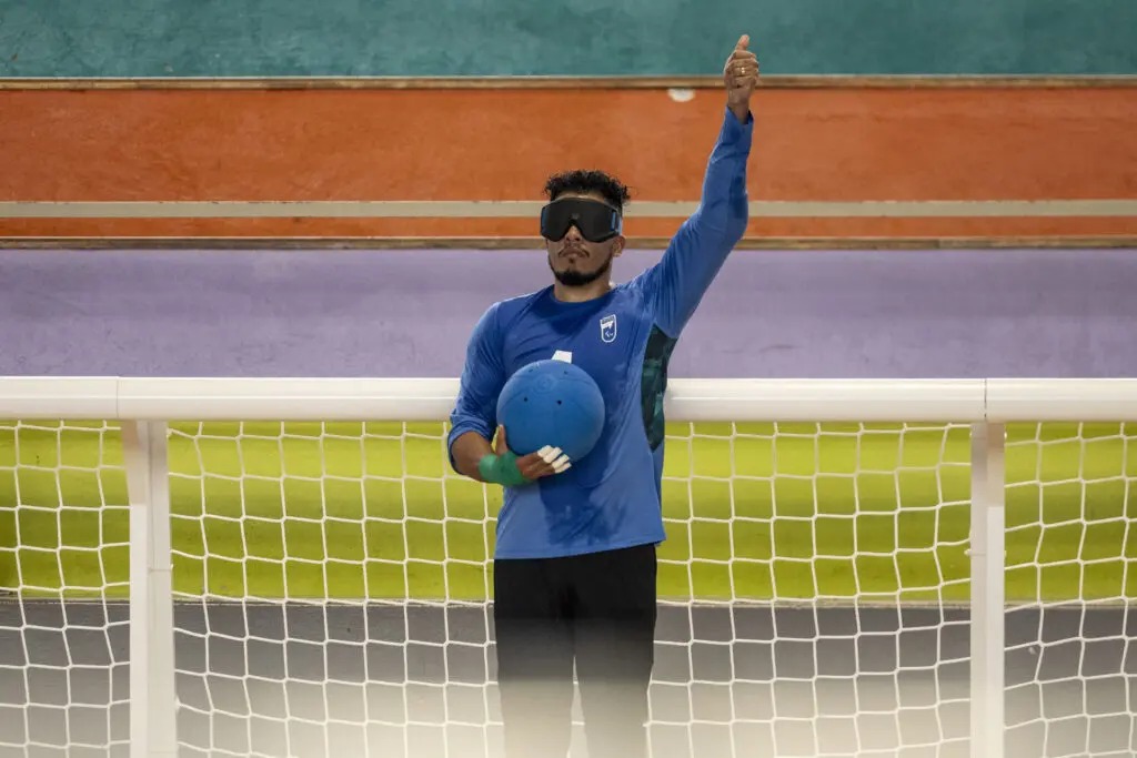 Leomon Moreno usa blusa de manga azul e se posiciona com uma bola na mao direita e a mão esquerda levantada em frente a um gol, em uma quadra de goalball 