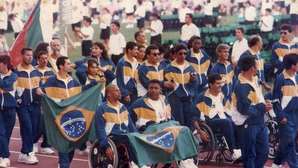 Varios atletas brasileiros com uniforme de frio amarelo com uma listra azul se posicionam em uma pista de atletismo.