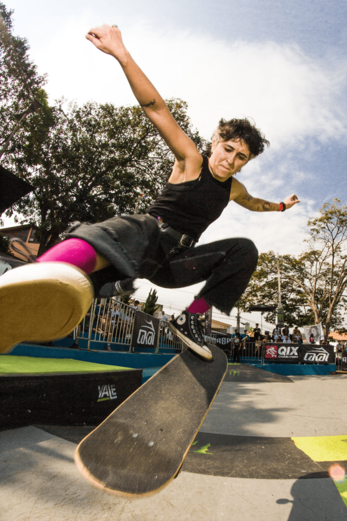 Atleta Feminina de Skate realizando manobra em uma competição oficial do esporte. Ela está usando uma camiseta e calça jeans preta e meias rosas.