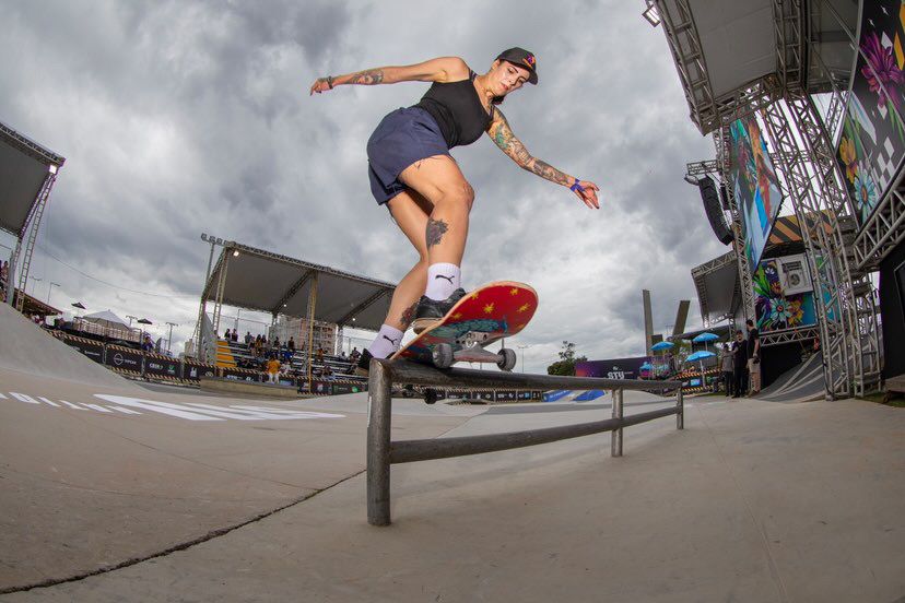 Atleta de skate de boné preto, blusa preta e short azul, meia branca e skate vermelho realizando manobra sobre corrimão em uma competição de skate.