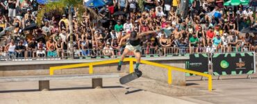 Atleta de skate de boné preto exercendo manobra sobre um corrimão amarelo