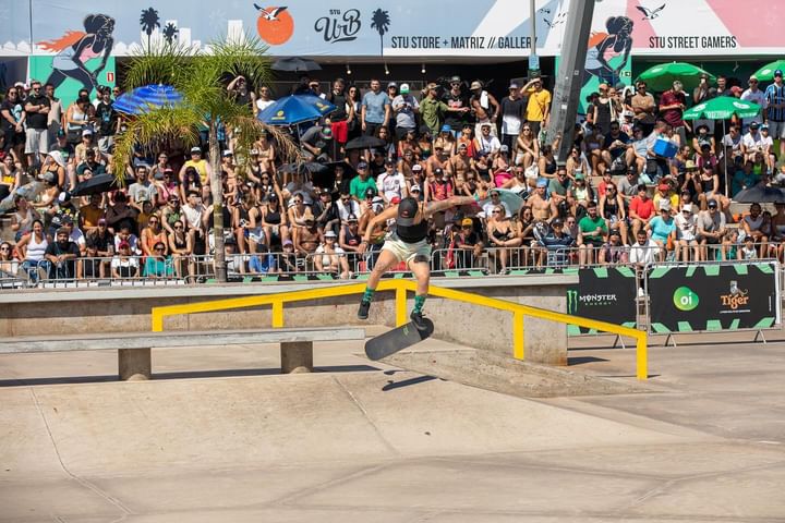 Atleta de skate de boné preto exercendo manobra sobre um corrimão amarelo