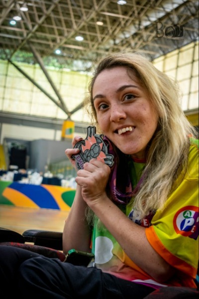 Amanda Alether, uma mulher branca e loira, sentada em uma cadeira de rodas, com um sorriso e uma medalha encostada no rosto