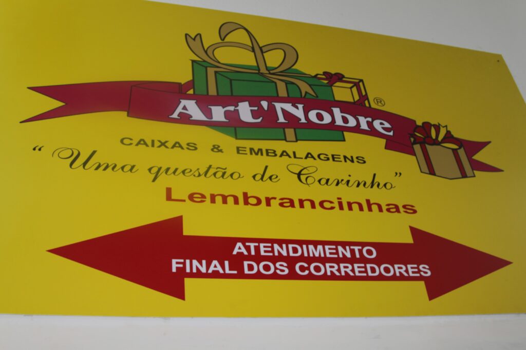 Placa amarela com a logo da loja "art nobre" que é composta por detalhes vermelhos, verdes e amarelos e alguns textos como "caixas e embalagens", "Uma questão de Carinho", "Lembrancinhas" e "Atendimento final nos corredores"