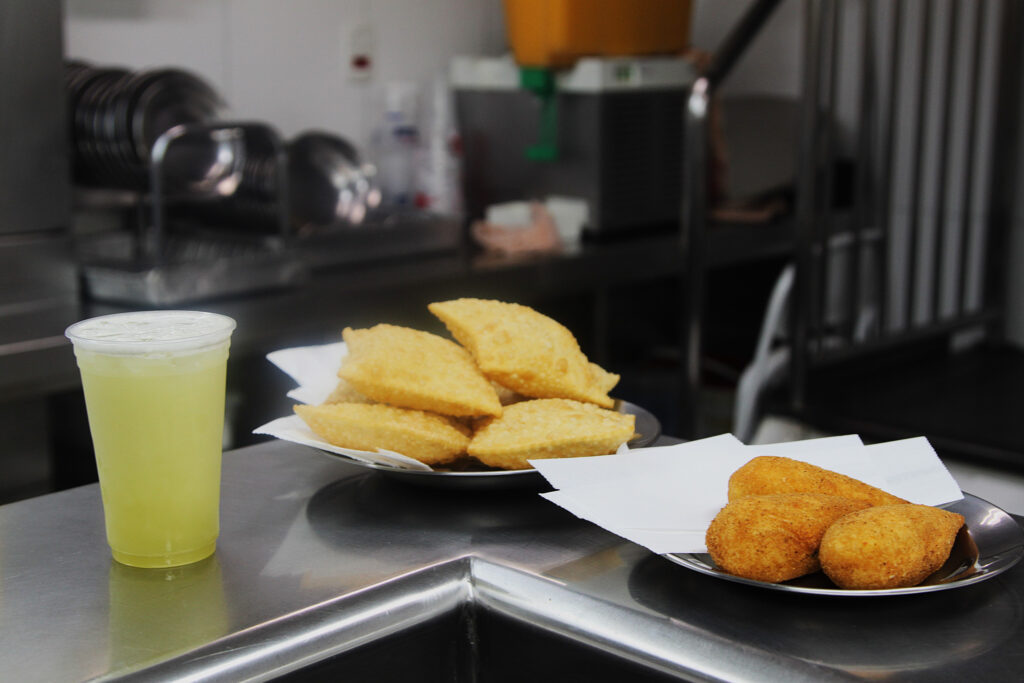 Imagem composta por um caldo de cana à esquerda, um amontoado de 4 pastéis em um prato ao centro e 3 coxinhas grandes em um prato à direita. O fundo é ocupado pela cozinha comercial da pastelaria desfocada