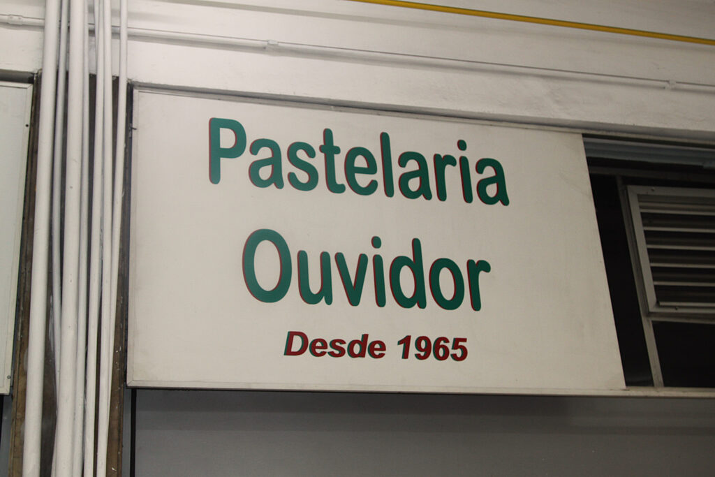 Placa da loja Pastelaria Ouvidor. A placa tem o fundo branco e é composta por apenas dois textos como elementos visuais: "Pastelaria Ouvidor" em verde e "Desde 1965" em vermelho