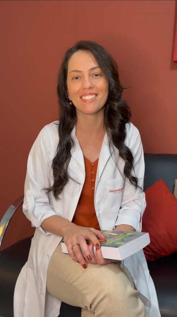 A nutricionista Alessandra Bertolino, uma mulher branca de cabelos logos, negros e ondulados, sentada com jaleco branco e blusa vermelha com um fundo vermelho, segura um livro de estudos da nutrição e senta-se de pernas cruzadas com uma calça nude.