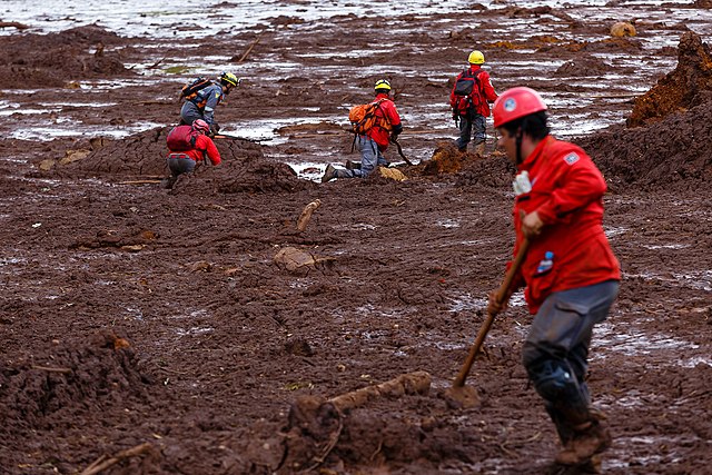 Bombeiros em uma área coberta por lama, realizando buscas e atividades de resgate. Eles seguram pás e outras ferramentas enquanto caminham e trabalham sobre a lama, que cobre o solo de maneira densa e irregular.