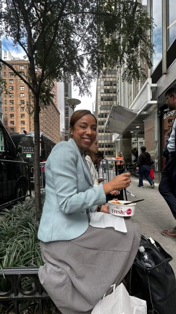 Edilene está sentada em um banco no na porta da Missão Brasileira em Nova Iorque, sorrindo e olhando na direção da câmera. Ela está de cabelo preso, usa brincos e veste um blazer azul claro que compõe com uma saia cinza. No momento ela está almoçando uma marmita fit e segura um garfo nas mãos, além disso há uma bolsa de couro preta do lado de seus pés.