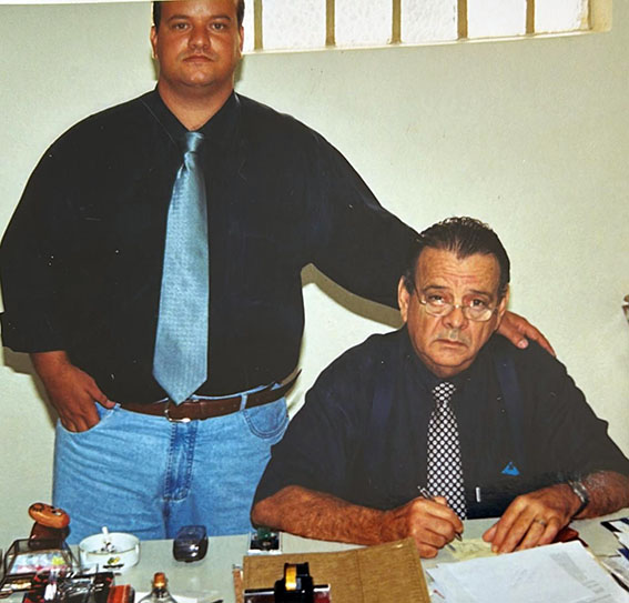 Ambos com camisa social preta e gravatas, Fernando Zuba com uma gravata cor azul claro e o Celso Zuba com uma gravata preta com bolinhas brancas. Celso sentado e apoiado em uma mesa com utensílios de escritórios, como canetas, cadernos, durex e papéis, como se estivesse escrevendo e Fernando em pé ao lado de seu pai.