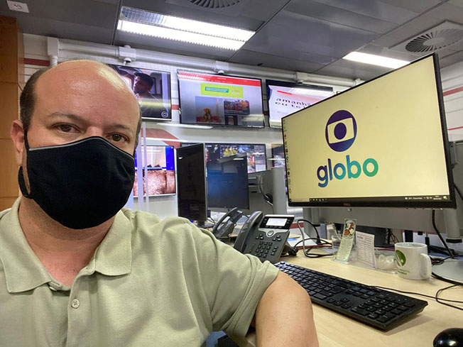 Fernando Zuba sentado em uma cadeira, apoiado em uma mesa com um telefone, xícara, teclado e um computador com a tela de fundo o símbolo da Globo. Utilizando máscara, aparente de seu rosto apenas seus olhos e sobrancelhas. 