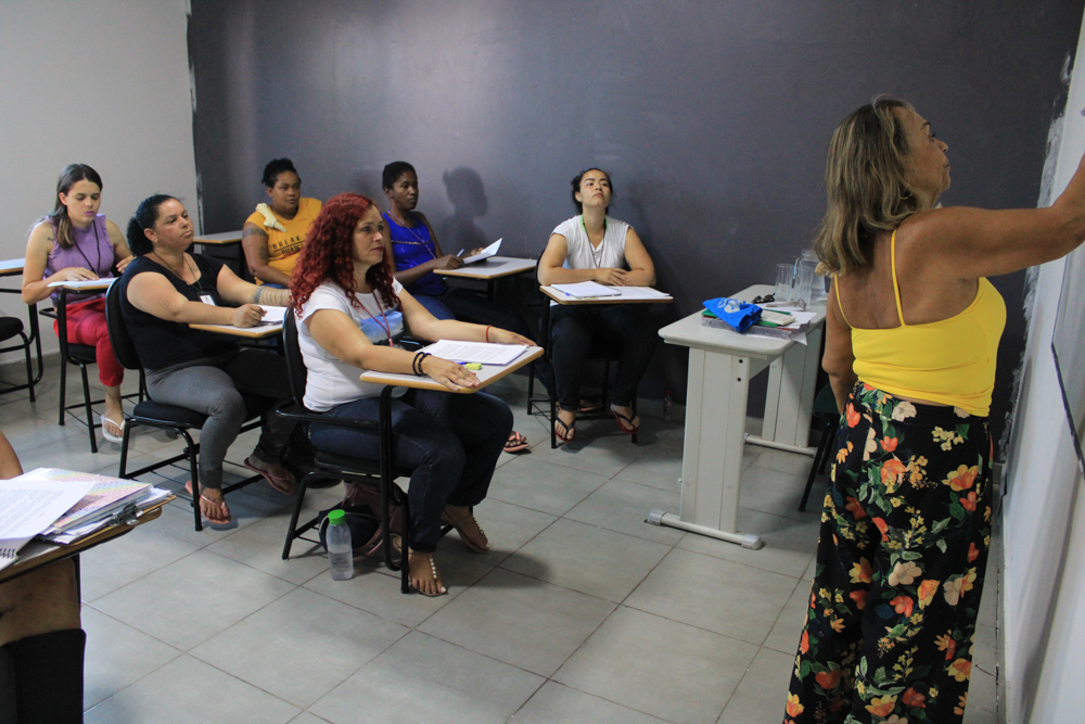 Sala de aula com uma professora na direita escrevendo na louça de costas para a turma que observa e compõe a parte esquerda da imagem