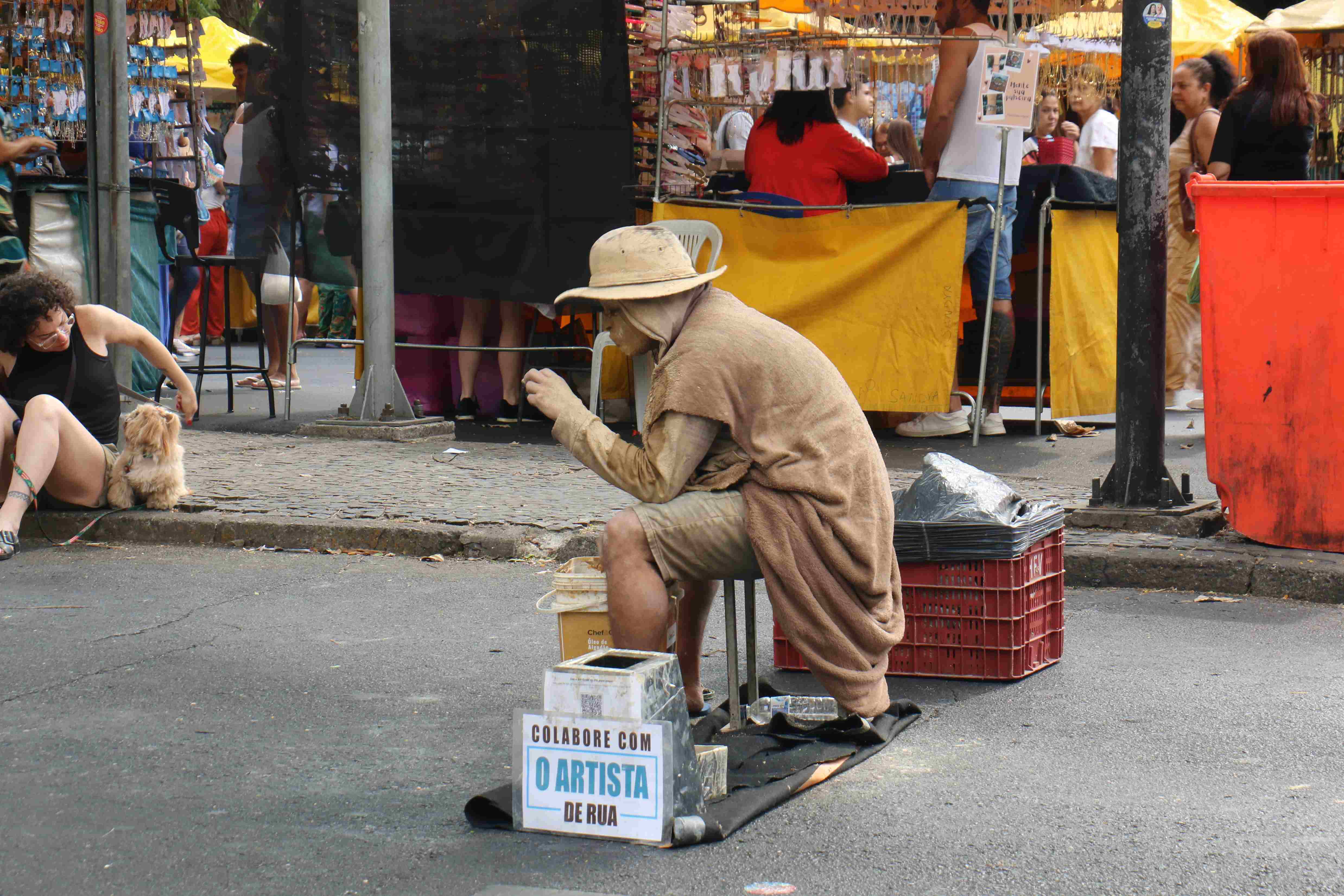 Artista que faz a performance de uma estatua viva coberta de "lama" e retocando a maquiagem, com uma placa de colabore com o artista de rua aos seus pés.