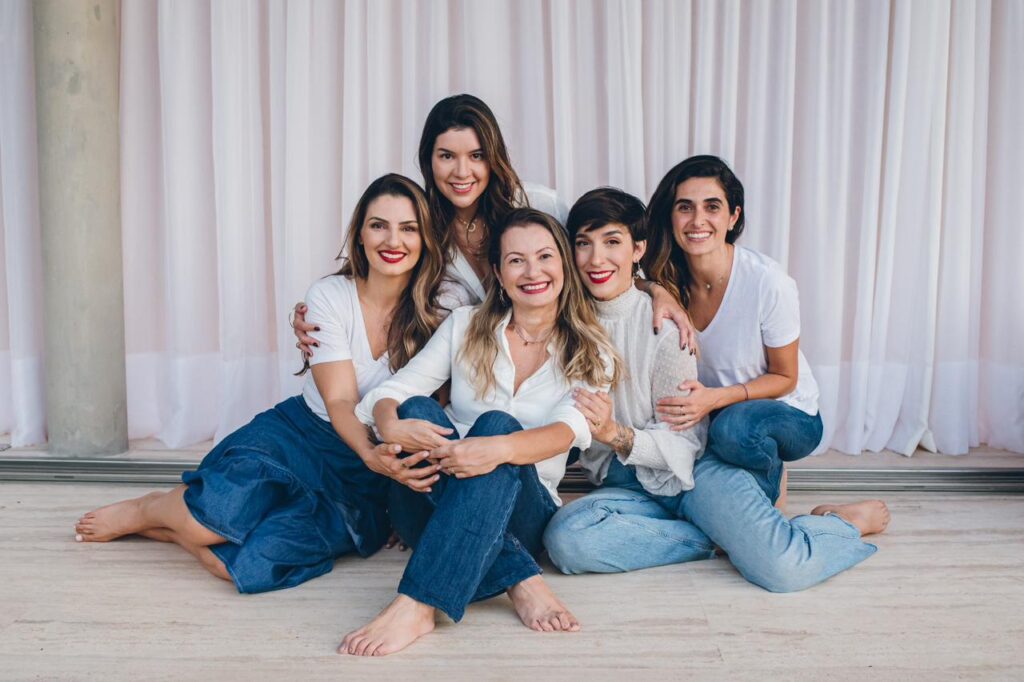 Mulheres sentadas juntas e se abraçando. Todas estão de blusas brancas e jeans.