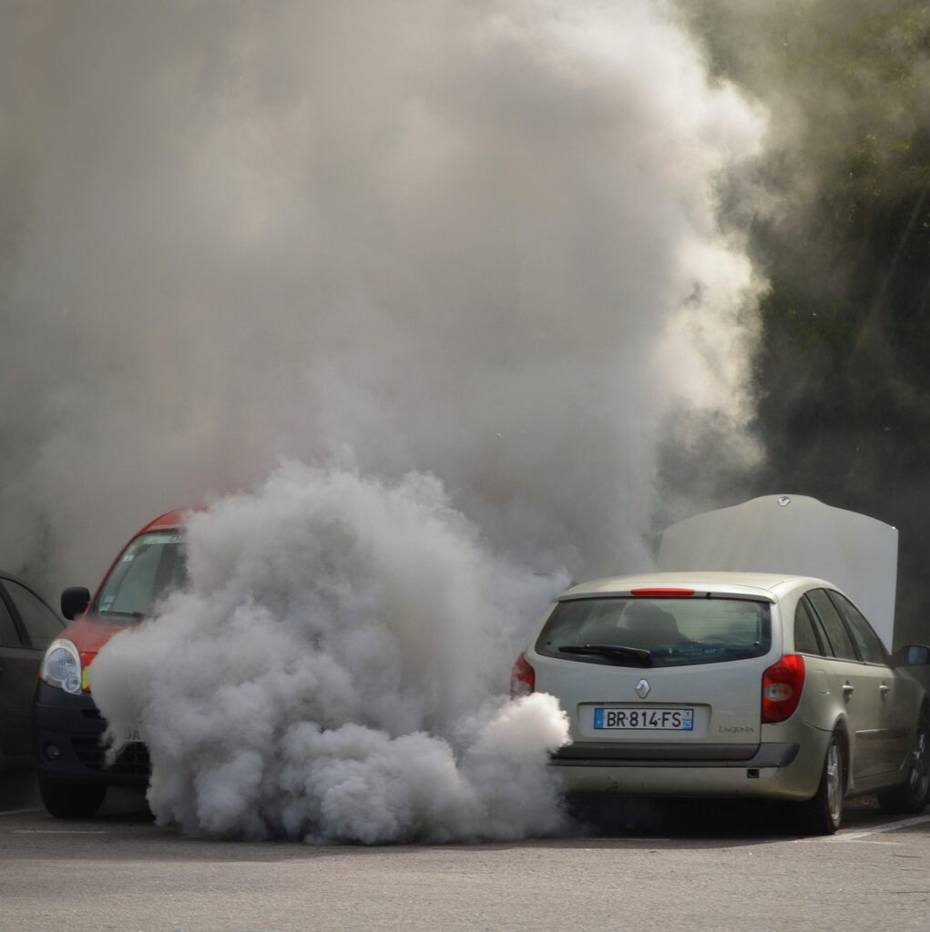 Dois carros estão parcialmente cobertos por uma grande nuvem de fumaça densa e cinza. O carro prateado está com o capô levantado, enquanto o veículo ao lado, de cor vermelha, também está envolto na fumaça que se espalha pelo ambiente, obscurecendo parte da cena. 