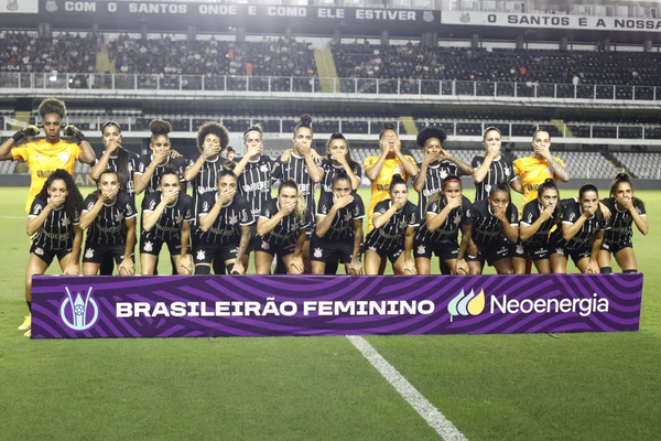 Jogadoras do Corinthians taparam a boca durante o Hino Nacional antes de jogo em protesto ao técnico do Santos Kleiton Lima. Time de jogadoras forma duas fileiras com a maioria em uniformes pretos com listras brancas e três com uniformes amarelos. Na altura de seus pés está faixa roxa com escritos em branco "Brasileirão feminino" 