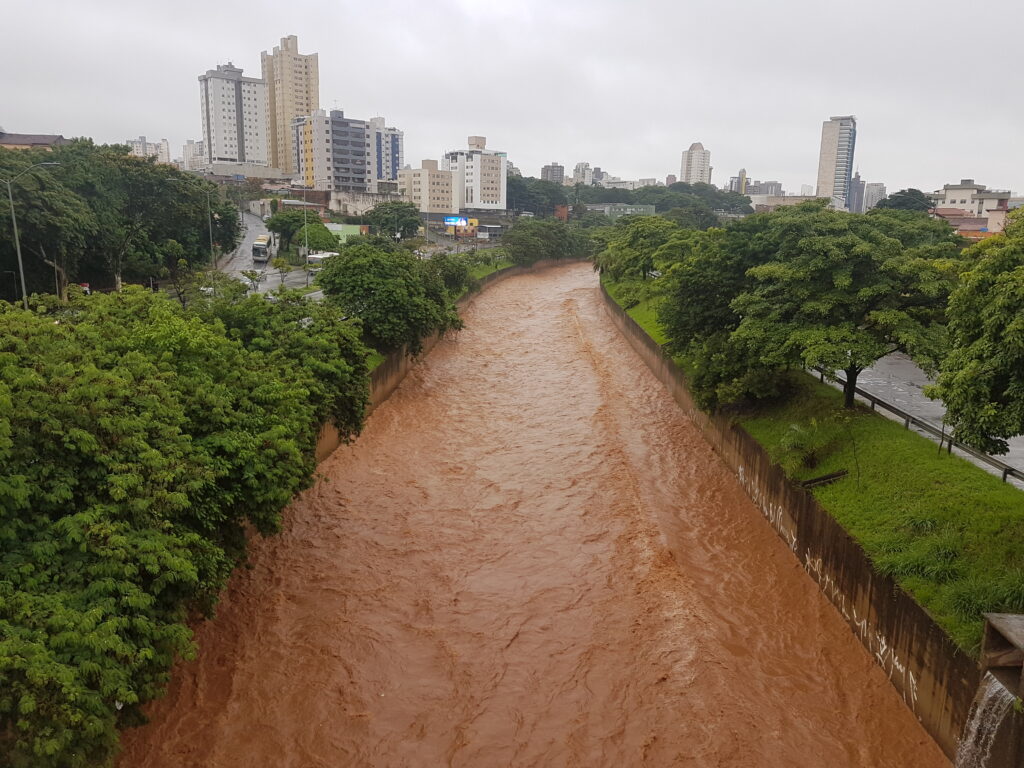 Curso d'água em Contagem, região metropolitana de BH com nível de agua elevado devido a chuvas. A cor da água é de um tom marrom e ao redor é possível ver vegetação. Ao fundo é possível  ver alguns prédios. 