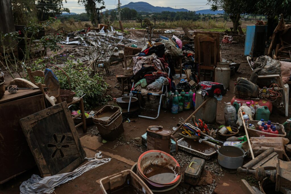 Inúmeros pertences pessoais como panelas, roupas, móveis completamente cobertos de lama do lado de fora de uma casa, e ao redor muitas árvores. 