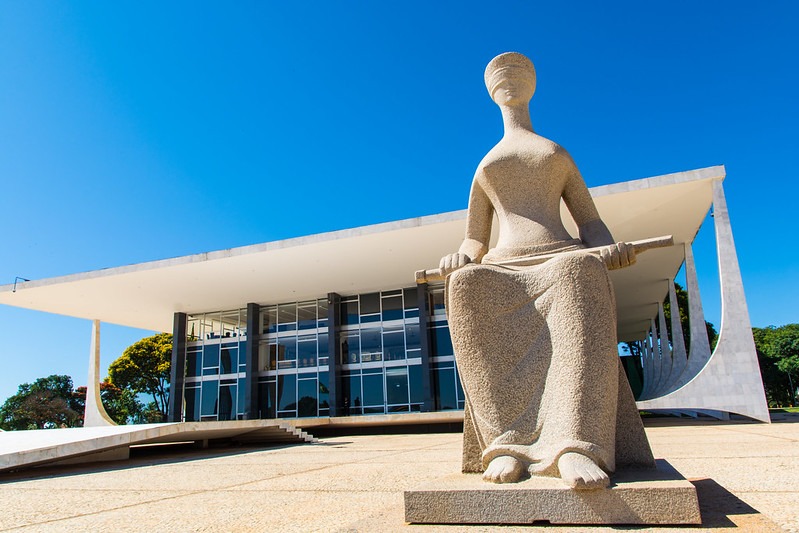 A imagem retrata a estátua da Justiça, localizada em frente ao edifício do Supremo Tribunal Federal (STF), em Brasília. A estátua, feita de pedra, representa uma figura feminina com os olhos vendados, simbolizando imparcialidade, e segurando uma espada, que representa a lei.