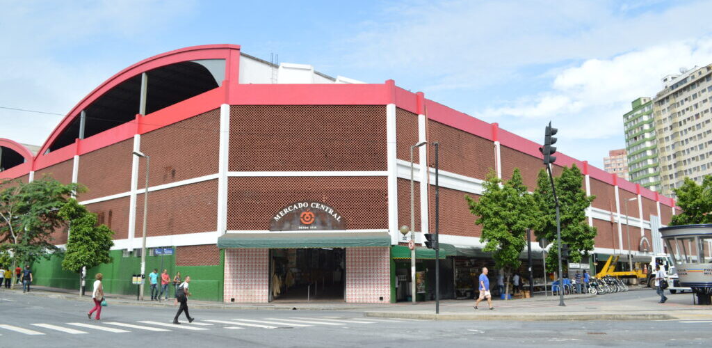 Fachada do Mercado Central de Belo Horizonte, um edifício com arquitetura tradicional de tijolos e detalhes em vermelho e branco. A imagem mostra o cruzamento de ruas em frente ao mercado, com pedestres e árvores ao redor, em um dia ensolarado.