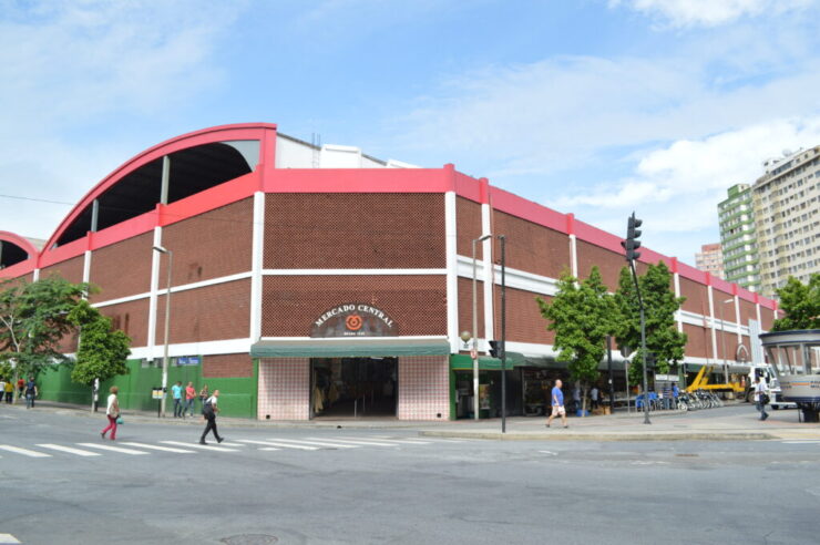 Fachada do Mercado Central de Belo Horizonte, um edifício com arquitetura tradicional de tijolos e detalhes em vermelho e branco. A imagem mostra o cruzamento de ruas em frente ao mercado, com pedestres e árvores ao redor, em um dia ensolarado.