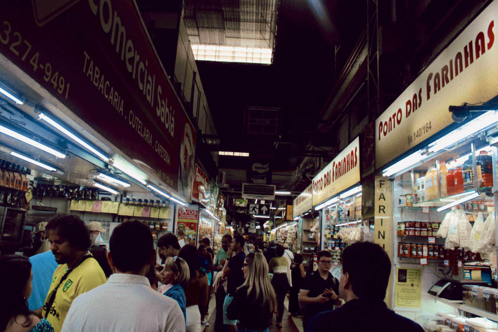 Corredor interno de um mercado com várias lojas e placas indicando estabelecimentos como 'Comercial Sabiá' e 'Ponto das Farinhas'. O local está movimentado, com pessoas caminhando e conversando entre os estandes, que exibem produtos como grãos, farinhas e itens diversos.