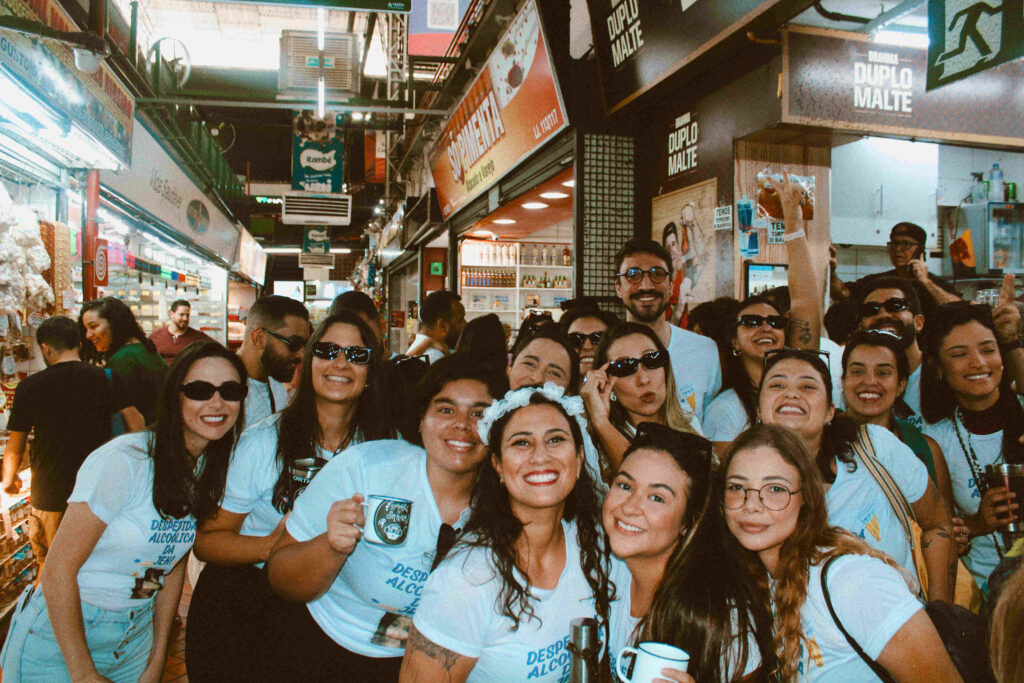 Grupo de pessoas sorrindo dentro de um mercado. Elas estão vestindo camisetas brancas personalizadas e aparentam estar celebrando um evento descontraído. O local apresenta estandes comerciais ao fundo, com diversas placas e produtos expostos.