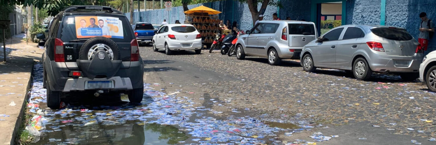 Uma rua mão única larga preenchida por carros parados em ambos os lados, à esquerda da foto um carro tem propaganda políica em seu vidro traseiro e, no chão da rua, muitos santinhos políticos