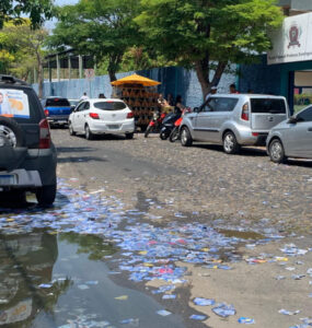 Uma rua mão única larga preenchida por carros parados em ambos os lados, à esquerda da foto um carro tem propaganda políica em seu vidro traseiro e, no chão da rua, muitos santinhos políticos