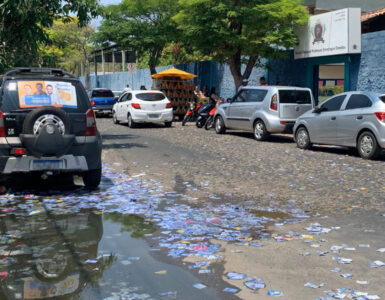 Uma rua mão única larga preenchida por carros parados em ambos os lados, à esquerda da foto um carro tem propaganda políica em seu vidro traseiro e, no chão da rua, muitos santinhos políticos