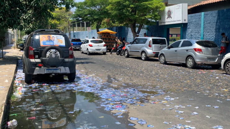 Uma rua mão única larga preenchida por carros parados em ambos os lados, à esquerda da foto um carro tem propaganda políica em seu vidro traseiro e, no chão da rua, muitos santinhos políticos