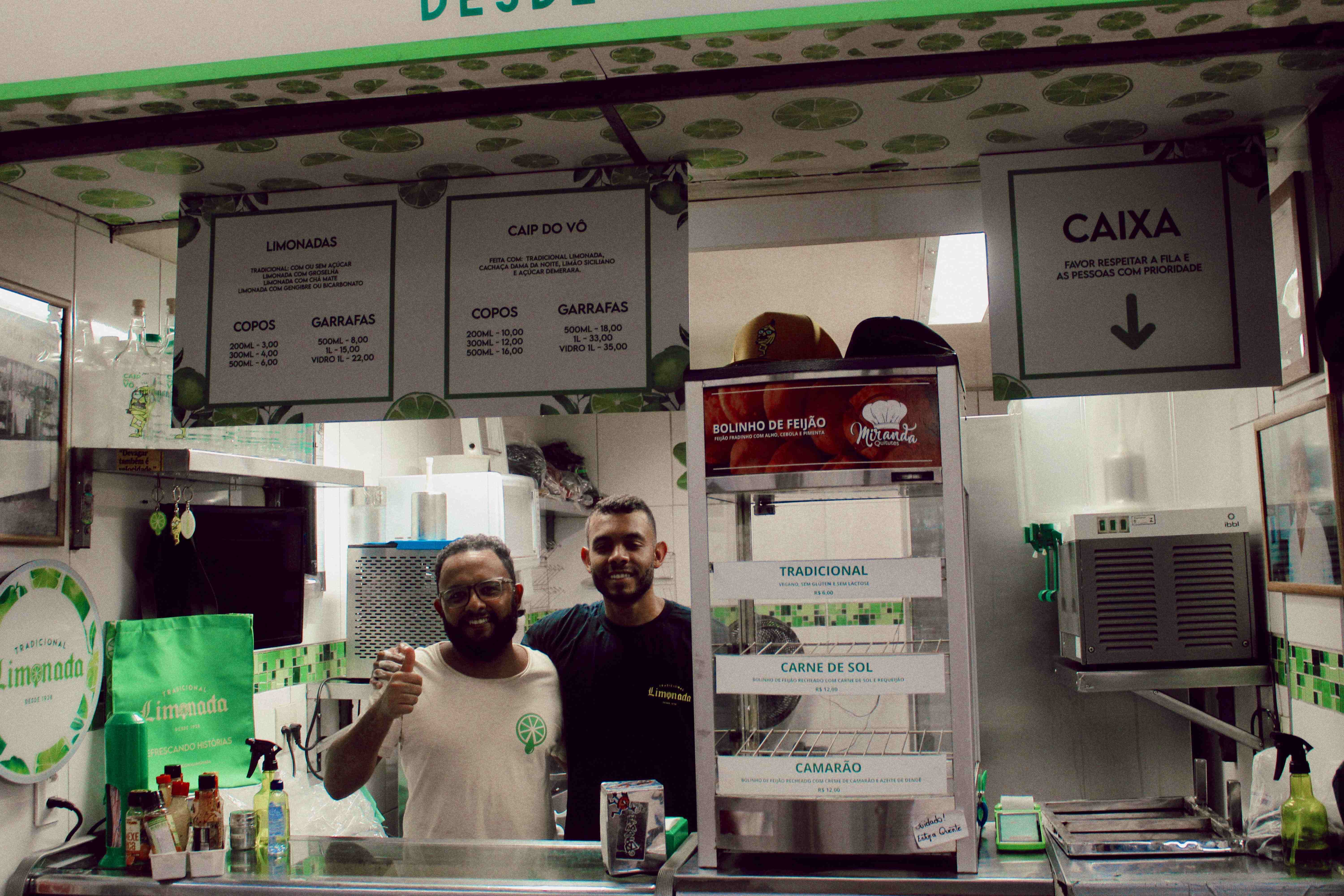 Dois atendentes sorrindo atrás do balcão de um estabelecimento, sendo um deles com óculos e camiseta clara com um logotipo de limão, e o outro com camiseta preta. O ambiente possui decoração temática de limão com cardápios detalhando bebidas como limonadas e 'Caip do Vô', além de uma vitrine exibindo opções de bolinhos de feijão. Ao lado direito, uma placa indica o local do caixa e equipamentos de cozinha completam o cenário.