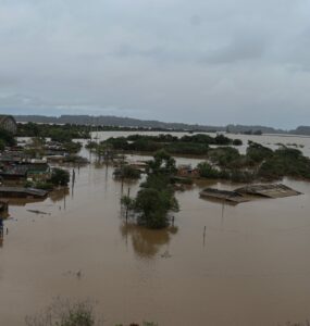 A imagem mostra uma área urbana alagada, com casas parcialmente submersas, postes e árvores emergindo da água. O tempo está nublado, indicando mais chuva.