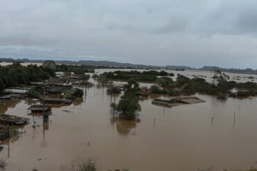 A imagem mostra uma área urbana alagada, com casas parcialmente submersas, postes e árvores emergindo da água. O tempo está nublado, indicando mais chuva.