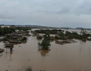 A imagem mostra uma área urbana alagada, com casas parcialmente submersas, postes e árvores emergindo da água. O tempo está nublado, indicando mais chuva.