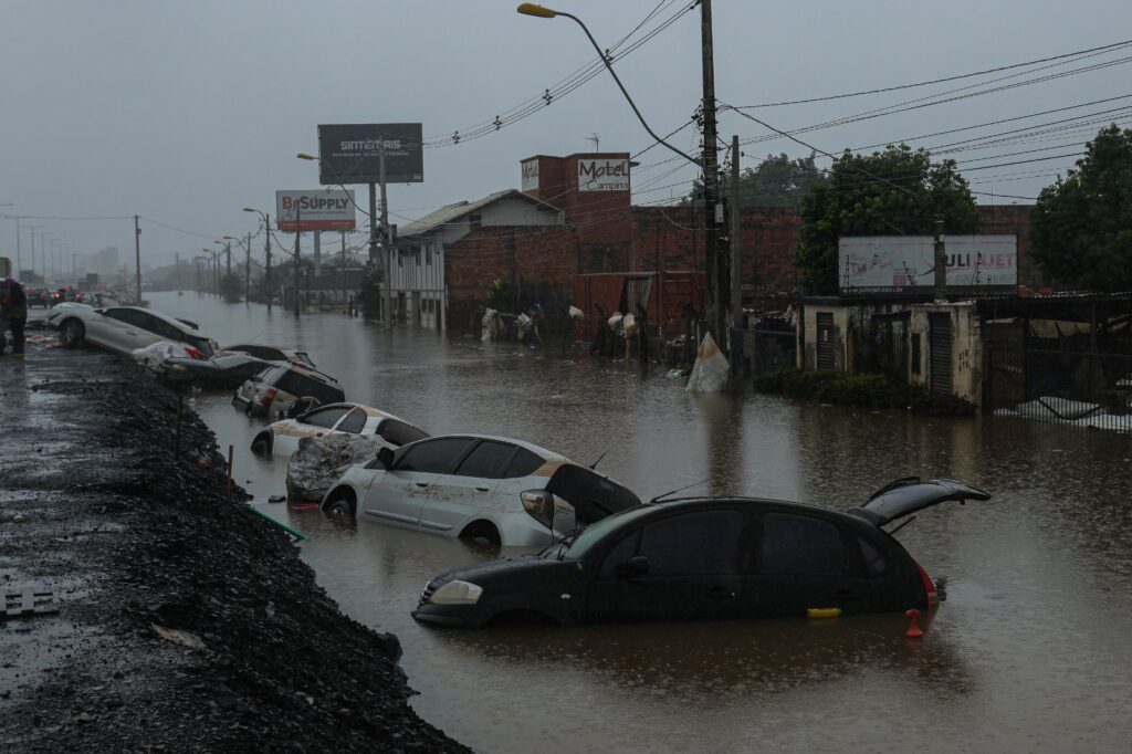 Avenida de um bairro completamente alagada, diversos carros parcialmente submersos na água que se elevou. O tempo está nublado. Ao redor dos carros é possível ver residências e postes também submersos.