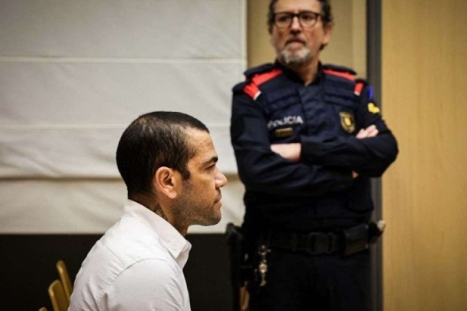 Daniel Alves sentado de perfil com roupa branca tatuagem a mostra e policial ao fundo em seu julgamento