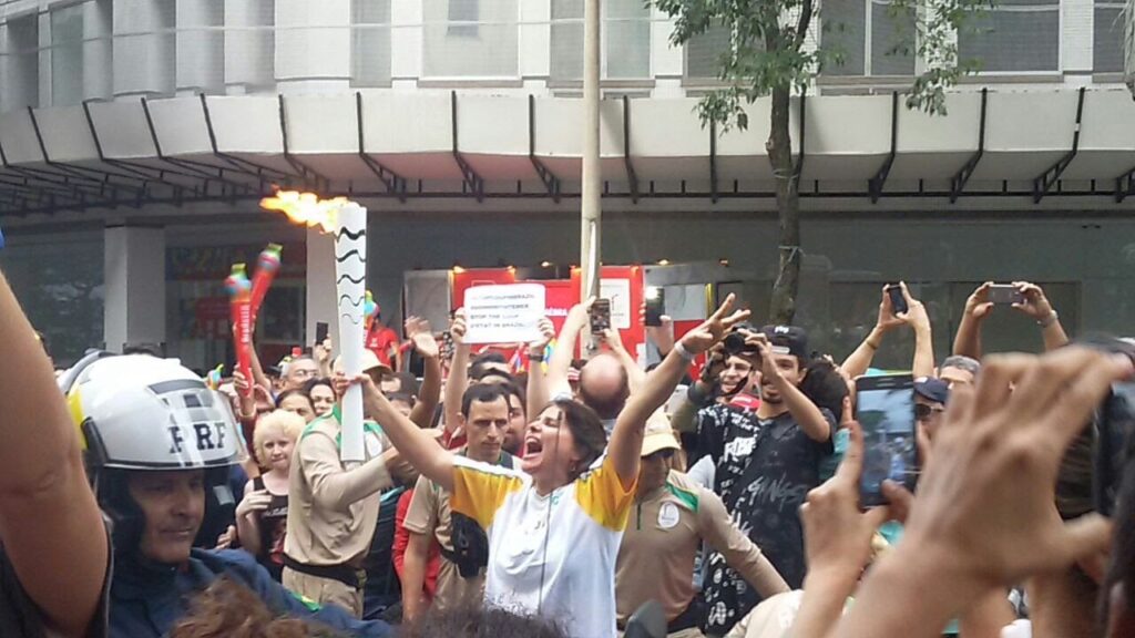 Mulher centralizada com os braços pra cima e levantando uma tocha olímpica acesa rodeada de pessoas em um local público.