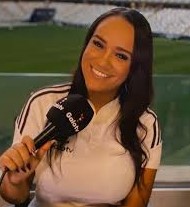 Carol Teixeira, uma mulher preta de cabelos longos e escuros, assim como seus olhos. Ela sorrindo, veste uma blusa branca e está segurando um microfone onde se lê Galo Tv com um gramado e arquibancadas ao fundo