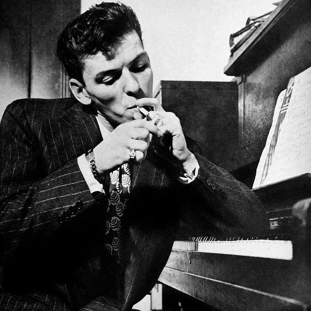 Fotografia em preto e branco. Apoiado em um piano, Frank Sinatra (jovem, branco, de cabelos castanhos e vestindo terno) acende um cigarro.