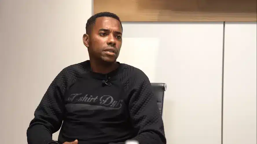 Jogador Robinho, um homem negro com cabelos curtos e escuros vestindo uma blusa de manga comprida preta sentado em uma cadeira dentro de um cômodo.