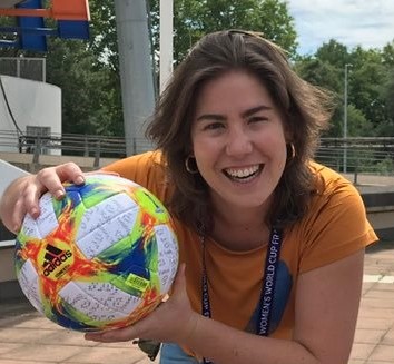 Maíra Nunes, uma mulher branca com cabelos curtos e castanhos e olhos castanhos. Ela está usando uma blusa laranja e segurando uma bola de futebol com uma paisagem ao ar livre no fundo