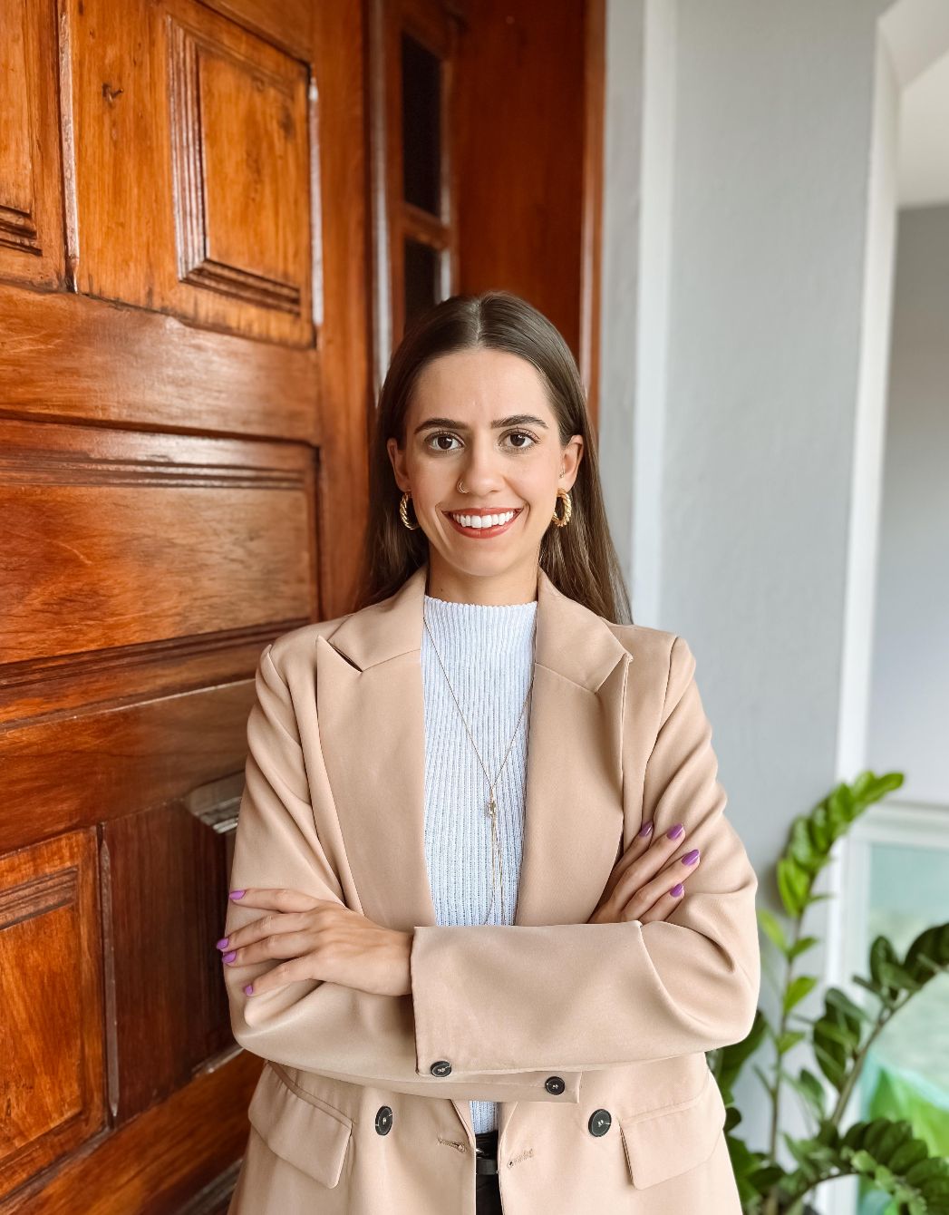 Mulher branca sorrindo e de braços cruzados, está usando um blazer bege e uma blusa branca. Encostada numa porta de madeira.