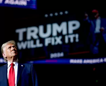 O presidente eleito dos Estados Unidos, Donald Trump, de pé, usando terno cor azul marinho e gravata vermelha, em frente a um telão escrito “Trump Will Fix It” (“Trump irá consertar tudo”, traduzido do inglês).